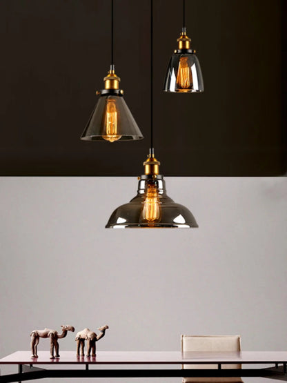 Three smoke grey glass pendant lights in cone, bell and dome shapes with brass fittings and glowing Edison filament bulbs against a dramatic dark and light split background above a minimalist dining table — LivDecor