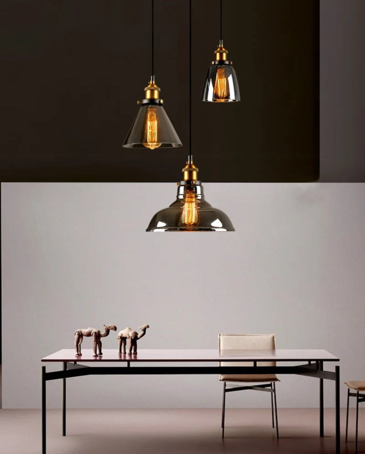 Three smoke grey glass pendant lights with brass fittings showing cone, bell, and dome shapes hung above a minimalist black dining table with camel figurines against a two-tone warm and dark wall — LivDecor