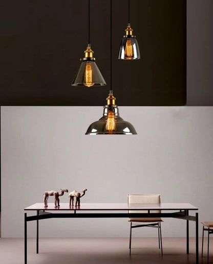 Three smoke grey glass pendant lights with brass fittings showing cone, bell, and dome shapes hung above a minimalist black dining table with camel figurines against a two-tone warm and dark wall — LivDecor