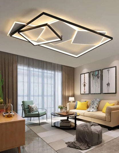 Strata Linear geometric LED chandelier in matte black glowing warm amber in a modern living room with cream sofa, yellow cushions, sage green armchair, and floor-to-ceiling curtains — LivDecor
