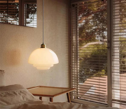 White glass dome pendant light with brass fitting illuminated above a bed with linen bedding and a low wood side table beside venetian blinds with a garden view — LivDecor