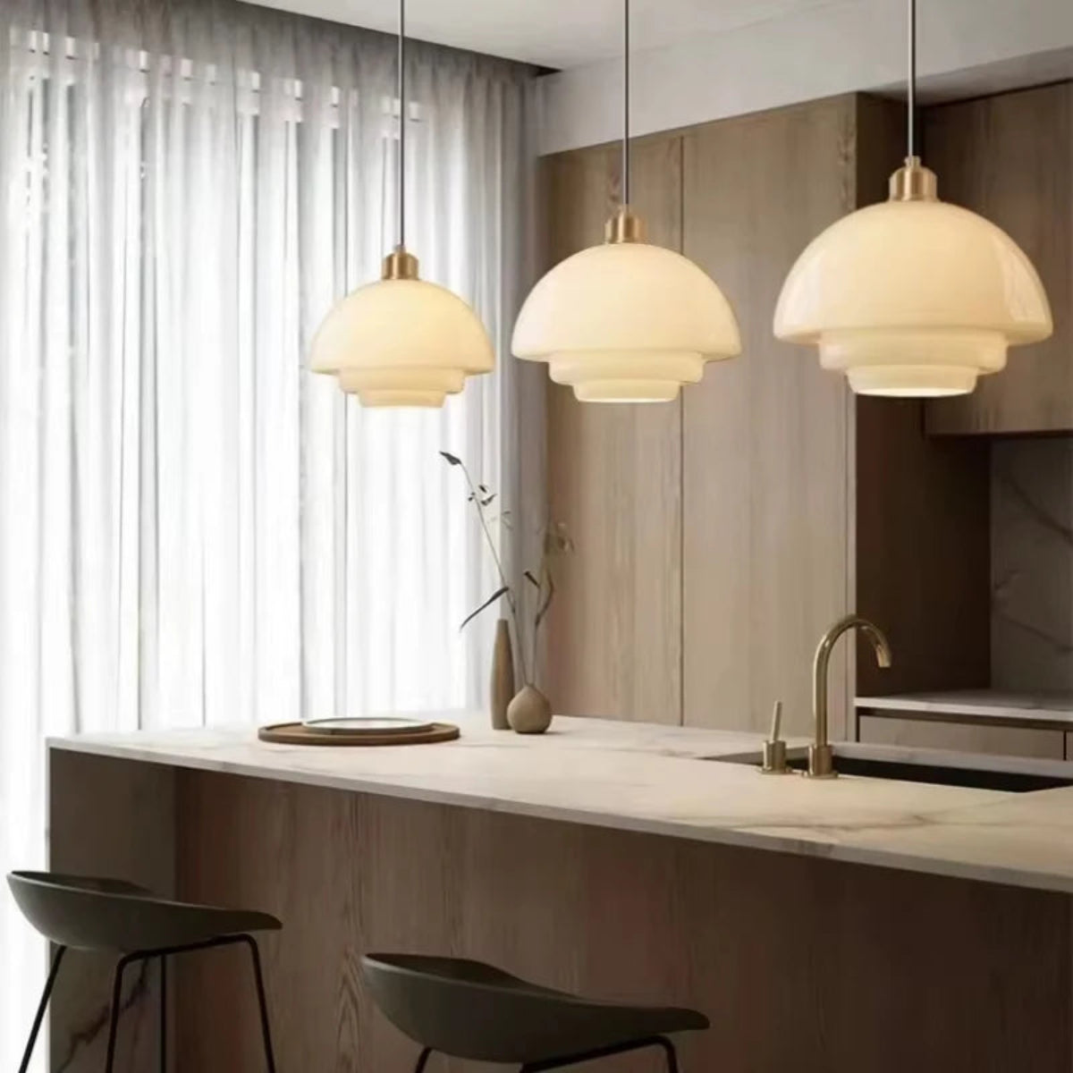 Three white glass tiered dome pendant lights with brass fittings hung above a marble kitchen island with warm wood cabinetry, sheer curtains, a ceramic vase, and brass tap — LivDecor