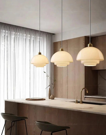 Three white glass dome pendant lights with brass fittings hung in a row above a marble kitchen island with warm wood cabinetry, sheer curtains, and bar stools — LivDecor