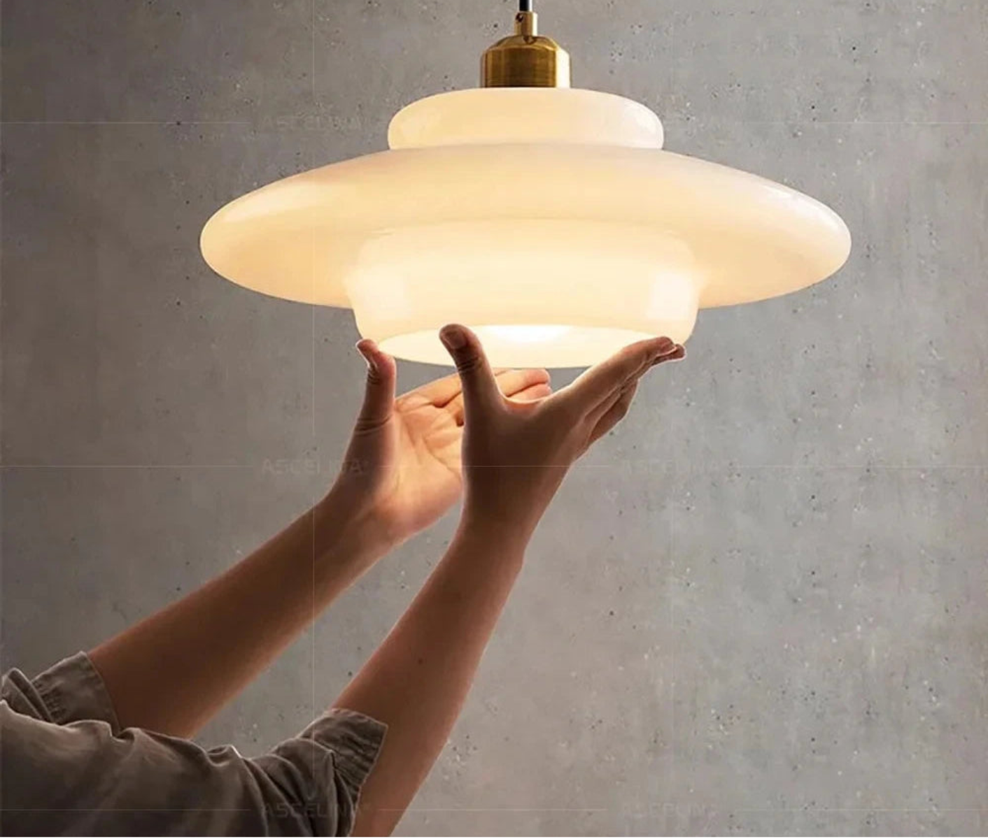 Hands holding a white glass saucer pendant light illuminated with warm light against a concrete textured wall, showing scale and warm glow — LivDecor